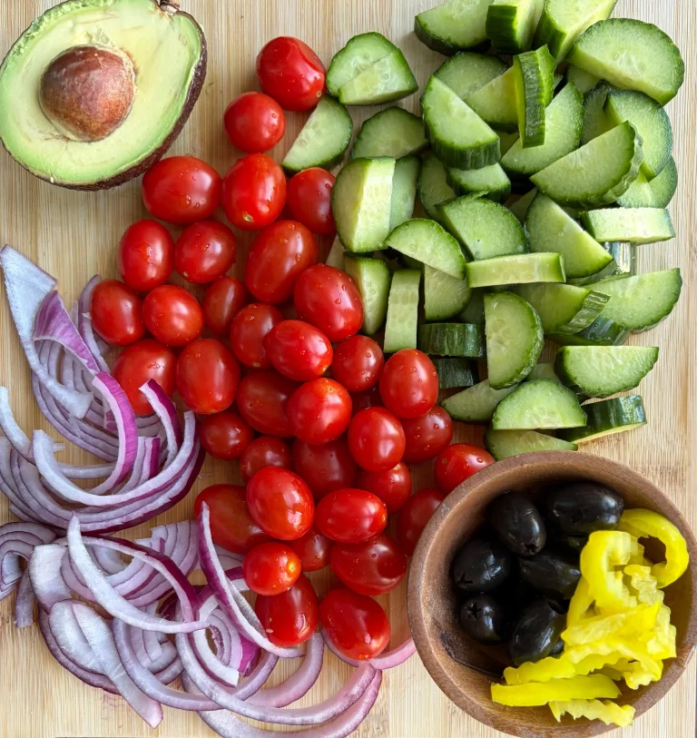 Sliced red onions, cherry tomatoes, avocado and cucumber on a wooden cutting board. Also black olives and banana peppers in a small wooden bowl.