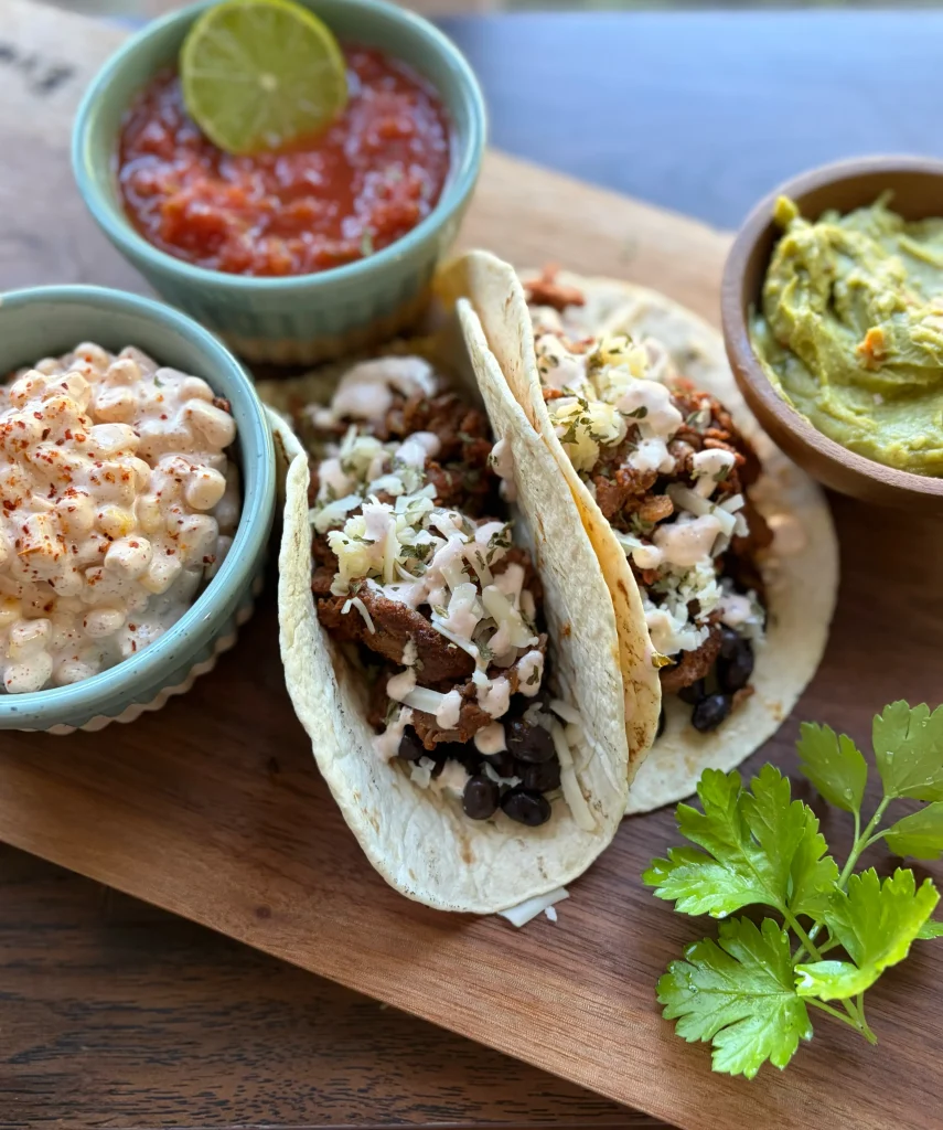 Steak tacos stacked next to each other. Corn dip, salsa, and guacamole on each side on top of a wooden cutting board.