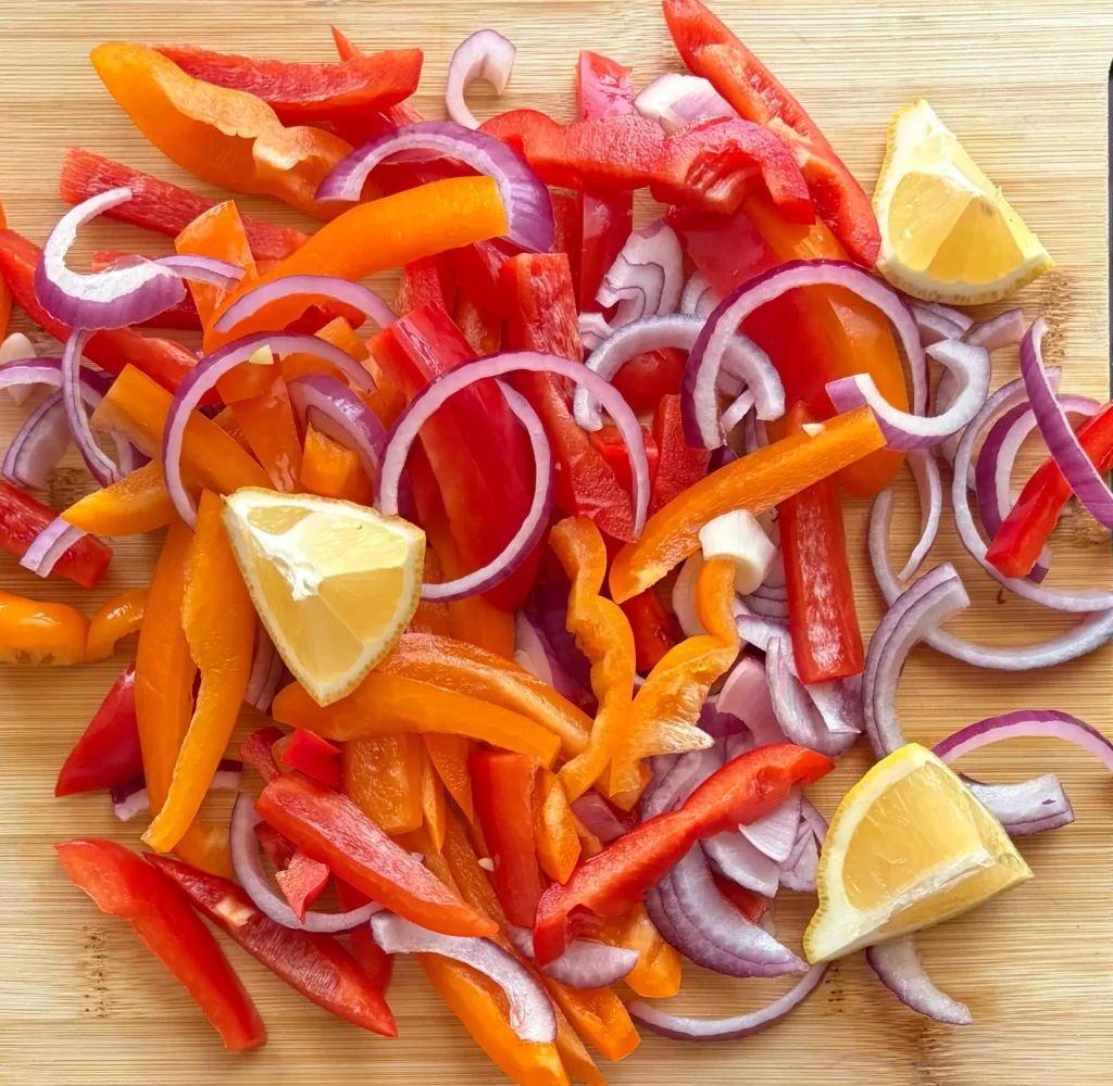 Red bell peppers, orange bell peppers, red onions, and lemon slices. Vegetables chopped on a wooden cutting board. 