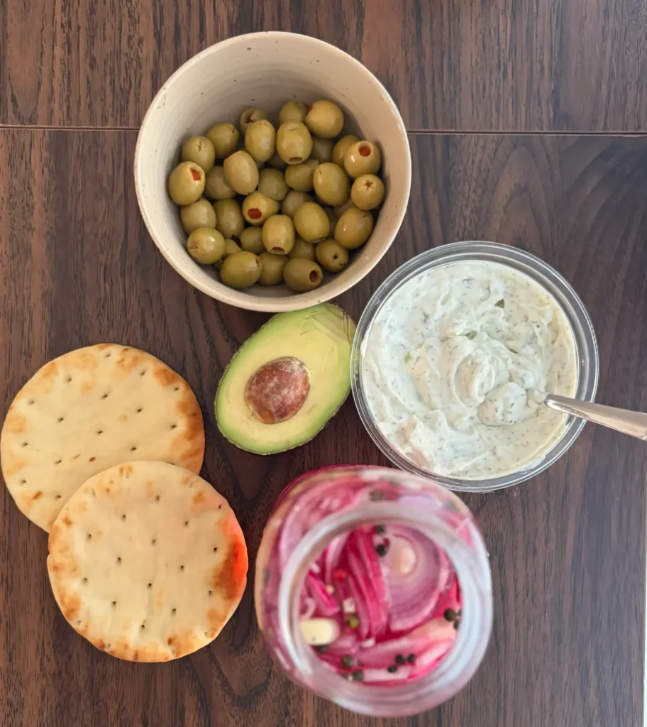 Green olives, pita bread, avocado, pickled red onions, and tzatziki sauce all lined up against a wooden background. 