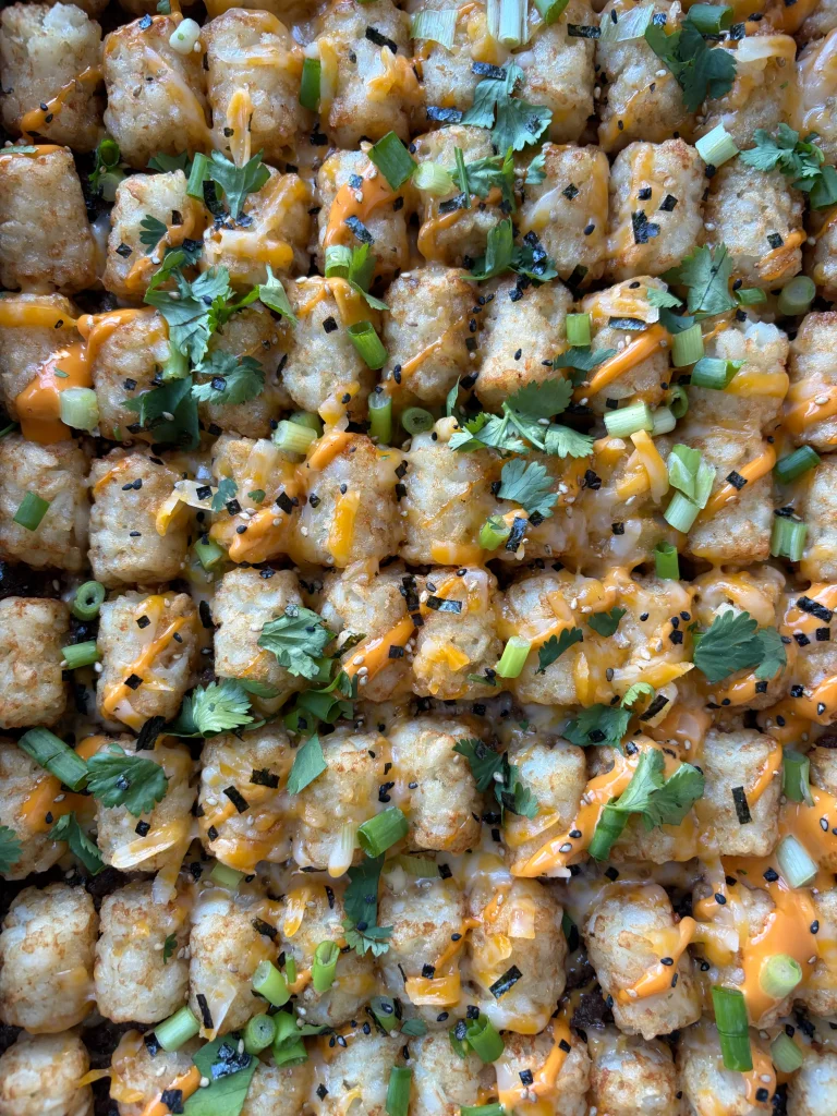 Tater tot casserole in a dish sprinkled with green onion and cilantro. Spicy mayonnaise drizzled on top.