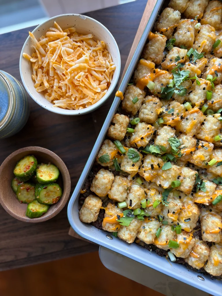 Tater tot casserole in a dish with cheddar cheese in a bowl and spicy cucumbers on the side.