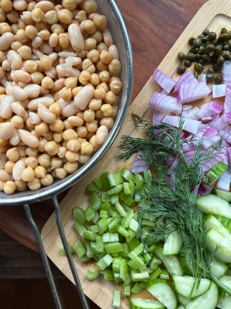 Drained chickpeas and white beans in a strainer next to a cutting board with celery, fresh dill, red onions, and cucumber.