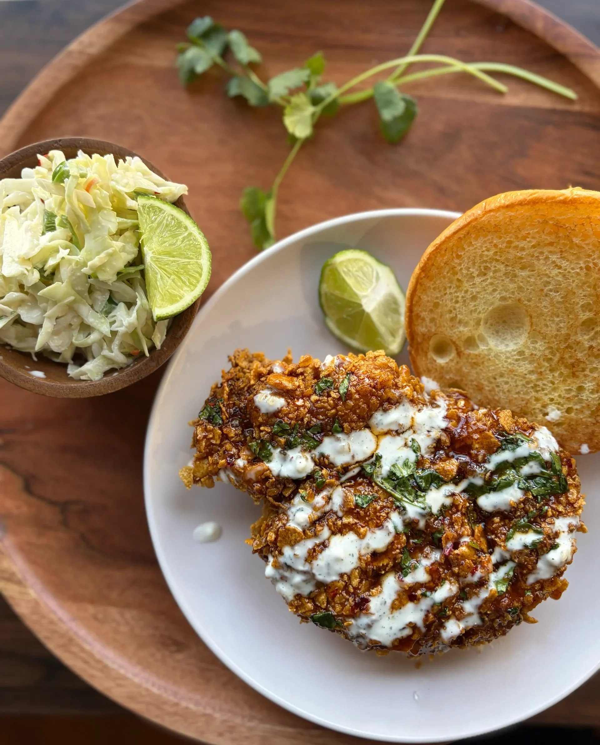Fried chicken sandwich with a ranch drizzle on top. A side of coleslaw with lime wedges and cilantro.