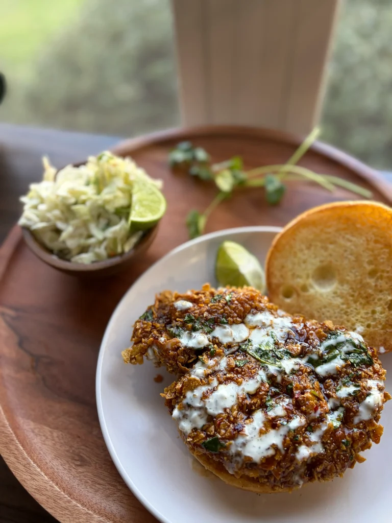 Fried chicken sandwich with a ranch drizzle on top. A side of coleslaw with lime wedges and cilantro.