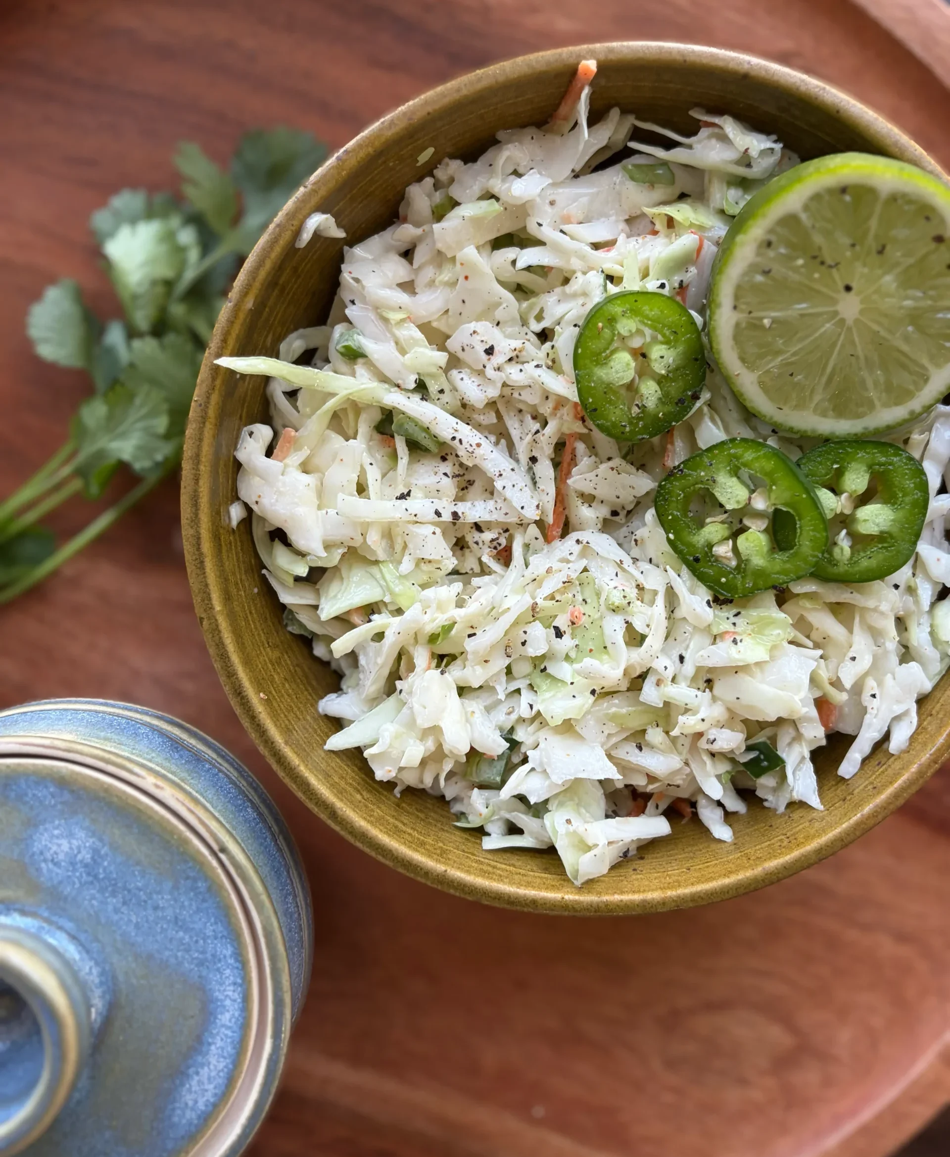 Creamy coleslaw with jalapenos slices and lime sitting on top in a bowl. A small blue container for decor.