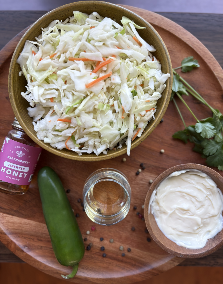 Coleslaw mix in a bowl, with mayonnaise and apple cider in a bowl on the side. A jar of honey and jalapeno laying on the cutting board.