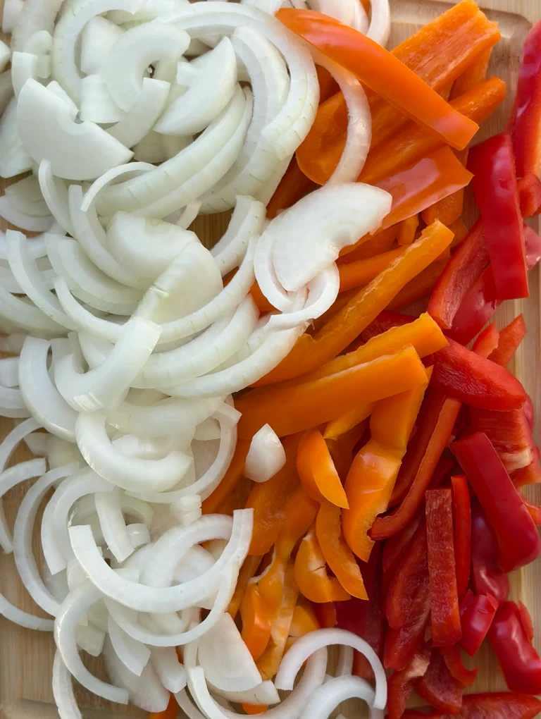 Sliced white onion, sliced orange bell pepper, and sliced red bell peppers on a cutting board.