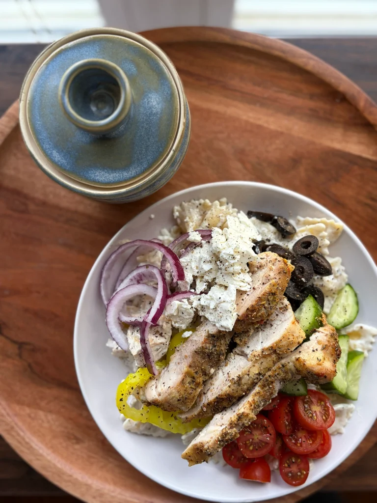 Pasta salad bowl topped with cherry tomatoes, cucumber, black olives, red onion, feta cheese, and grilled chicken thighs.
