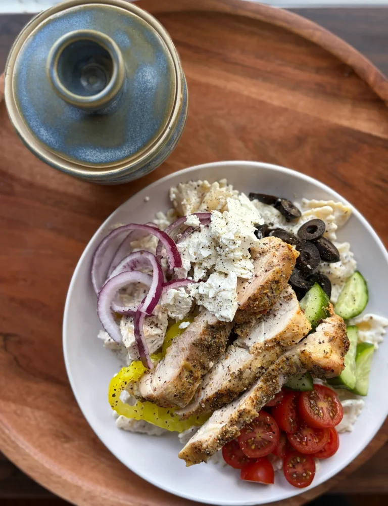 Greek pasta salad topped with grilled chicken, cherry tomatoes, red onion, and cucumbers. Small blue bowl for decoration.