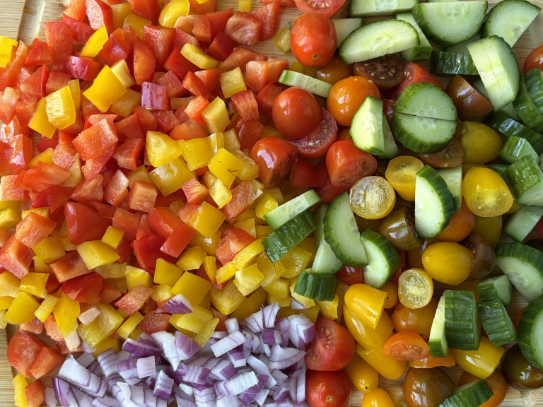 Chopped bell peppers, diced red onion, and diced cherry tomatoes, diced cucumbers. All arranged on a cutting board.