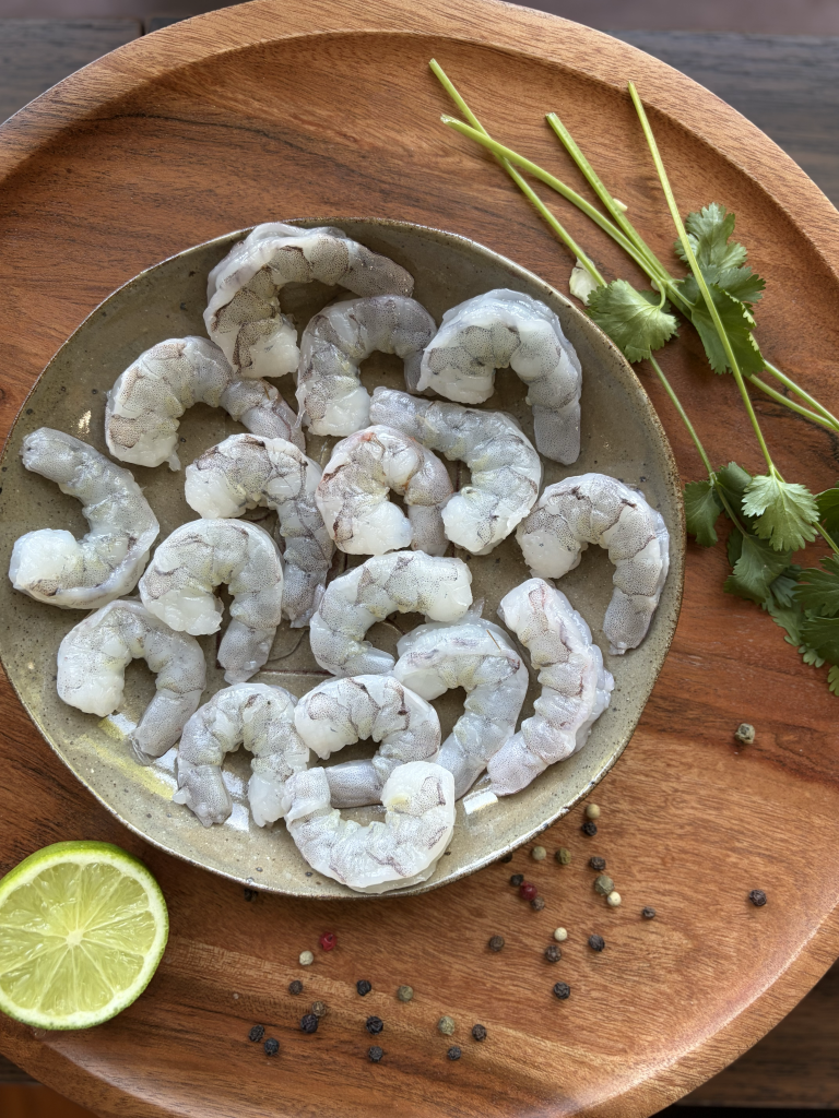 Raw shrimp in a bowl with a lime wedge and cilantro sprigs on the side.