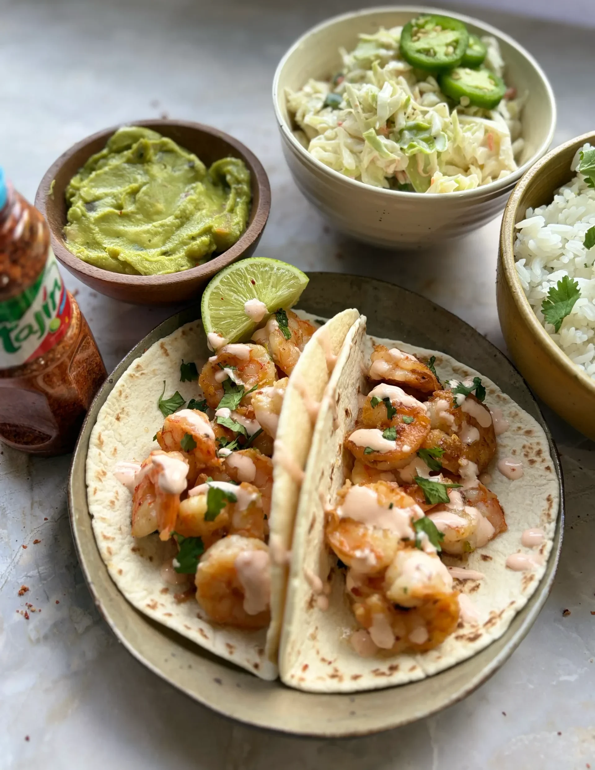 A plate of shrimp tacos with a bowl of guacamole and cole slaw on the side. There is a bowl of white rice to the right of the tacos.