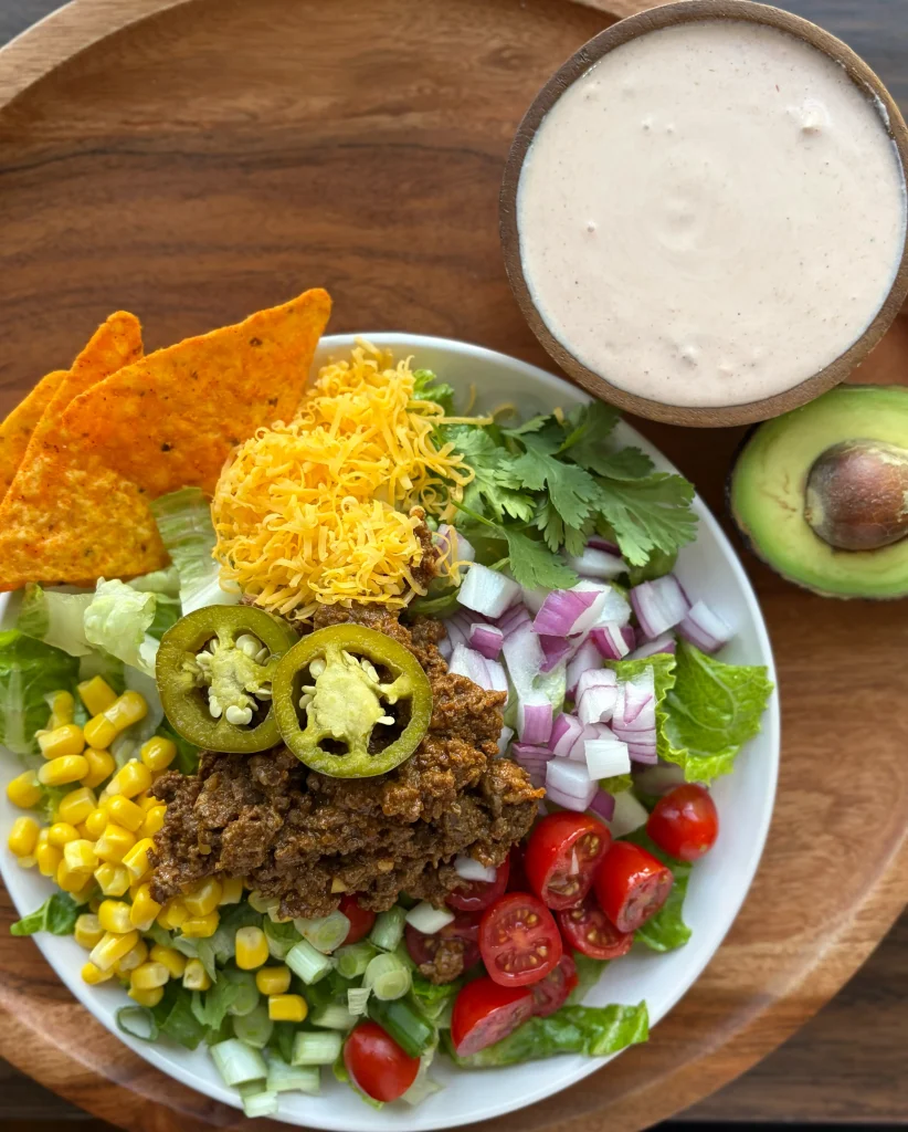 Taco salad with doritos and a creamy salsa dressing on the side. Also a sliced avocado in half on a wooden cutting board. 