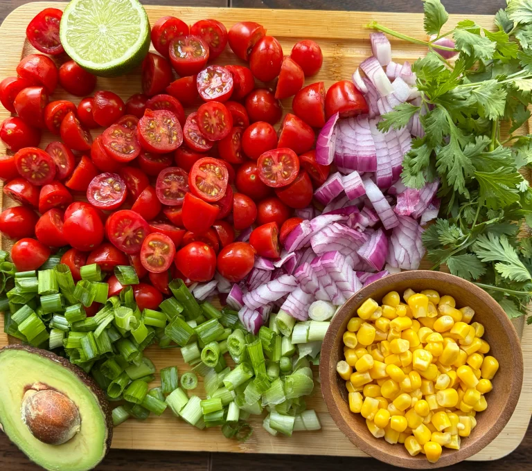 Sliced cherry tomatoes, diced red onion, diced green onion and a smlal bowl of corn on a wooden cutting board. Also half of an avocado and fresh lime for garnish.