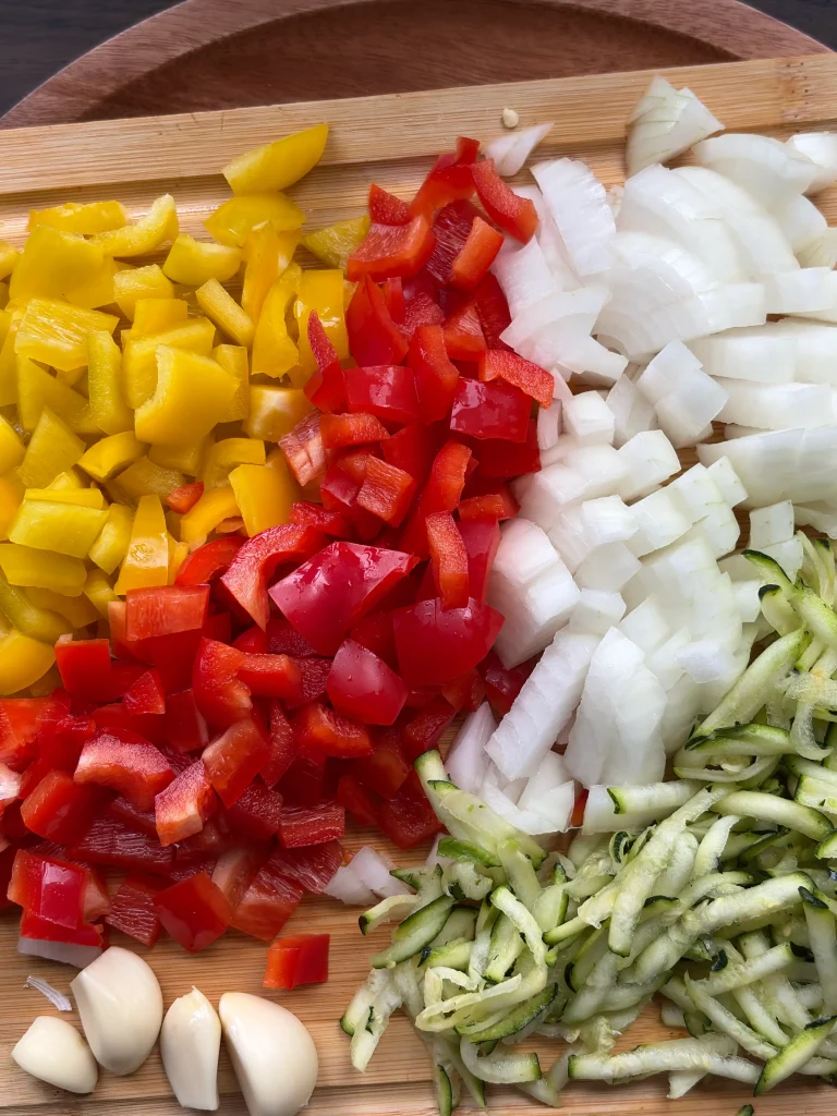 Diced yellow bell peppers, diced red bell pepper, diced white onion and grated zucchini on a wooden cutting board.