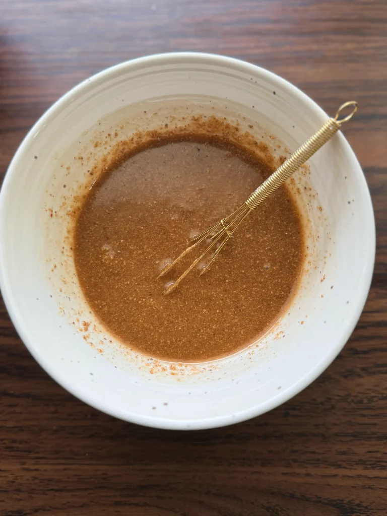 Bowl with almond butter and honey mixed with a golden whisk. All sitting in a white bowl.
