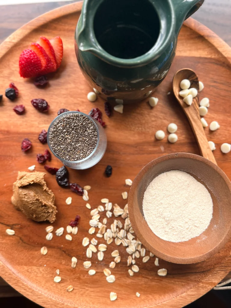 A wooden bowl with vanilla protein powder, a glass jar with chia seeds, and green pitcher for decor. Almond butter, dried berries, oatmeal, and white chocolate chips on a cutting board sprinkled around. 