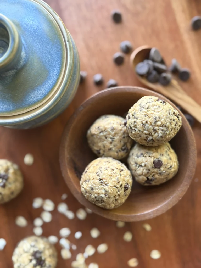 Cookie dough protein balls on a wooden bowl with chocolate chips on the side in a wooden spoon. A blue jar is on the side for decor.