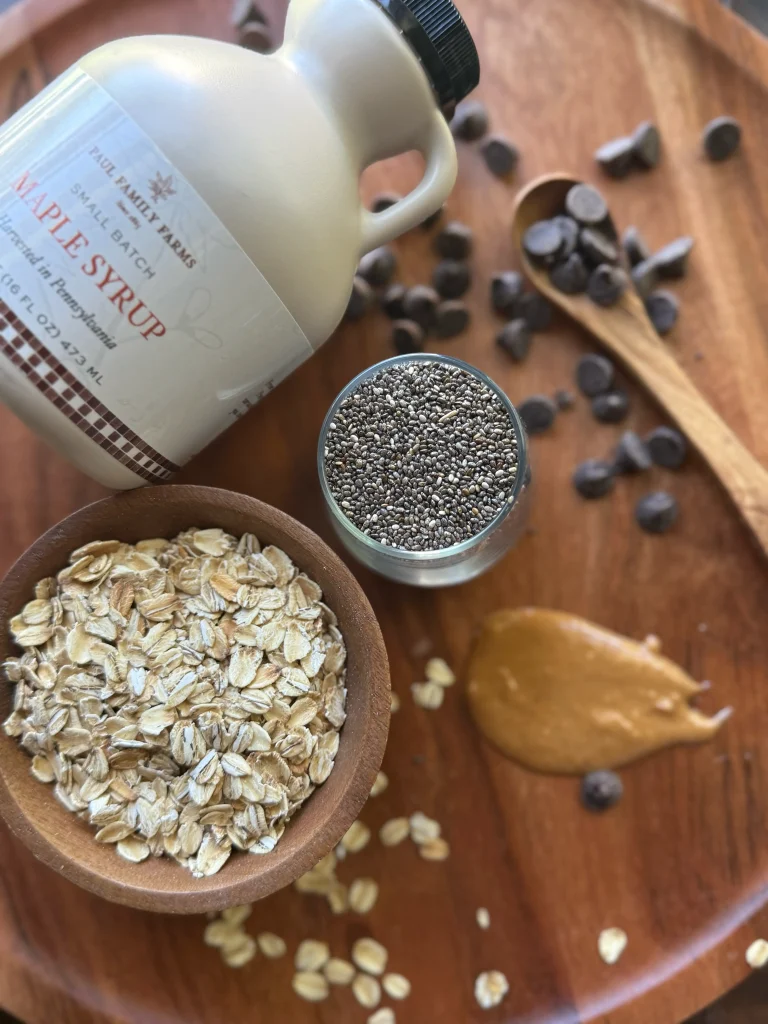 A bowl of uncooked oatmeal, a bowl of chia seeds, and a bottle of maple syrup on a wooden cutting board. Peanut butter and chocolate chips are sprinkled for decor.