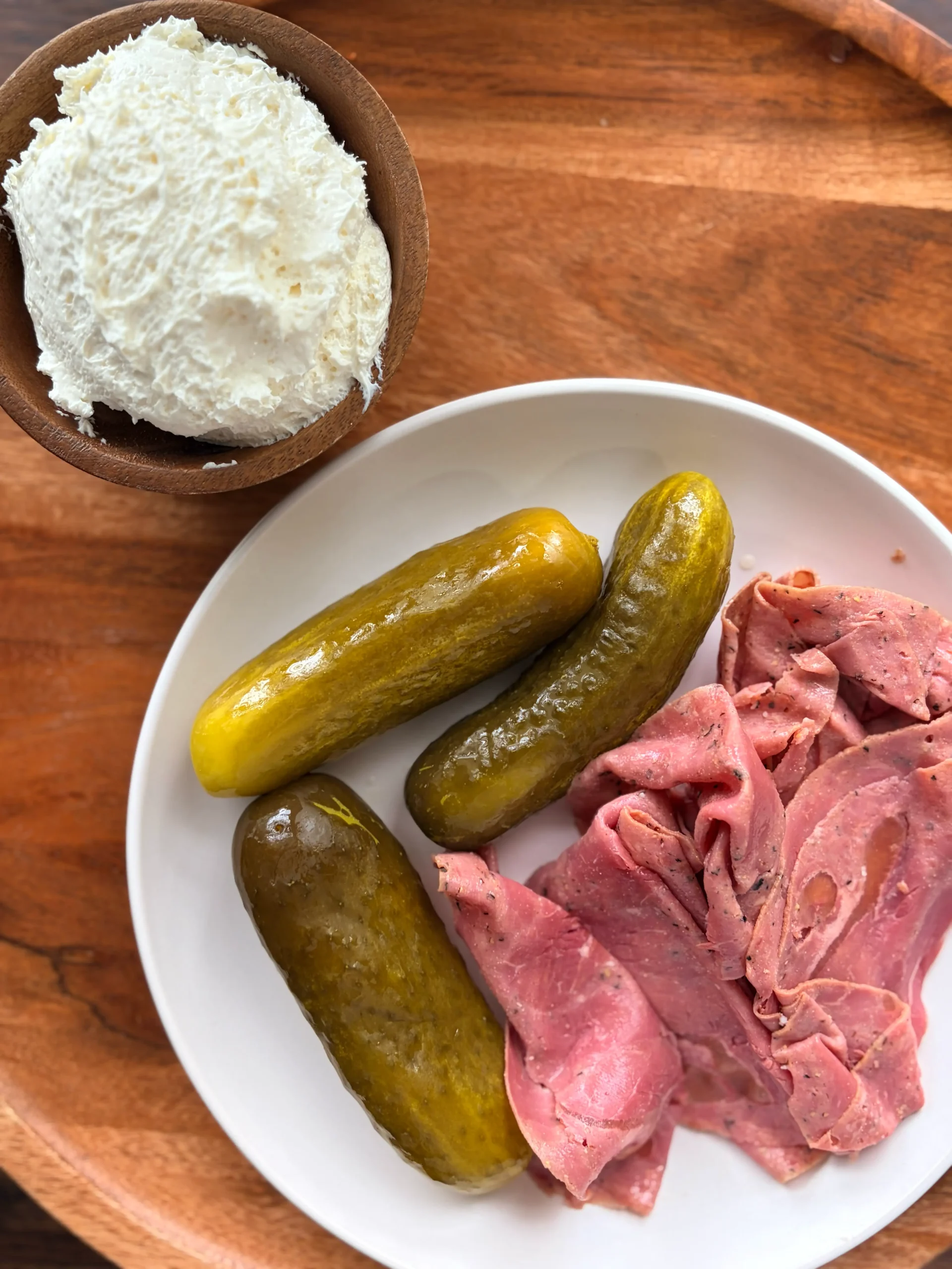 A plate with pastrami and pickles. A wooden bowl with whipped cream cheese is on the side.