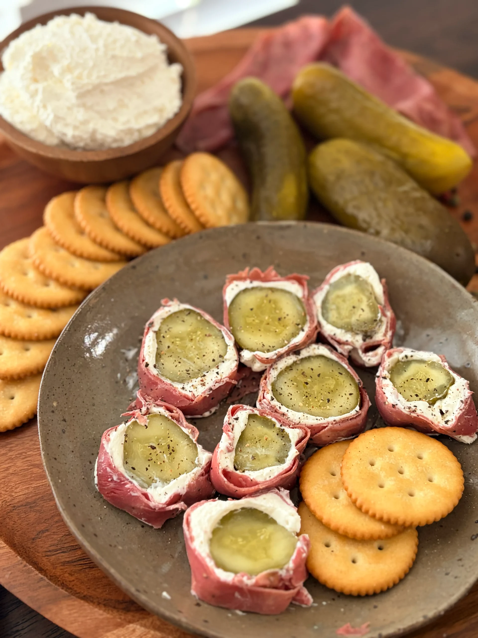 Pickle, pastrami, and cream cheese wrapped together and sliced on a plate. Ritz crackers and pickles are on the side for decor.
