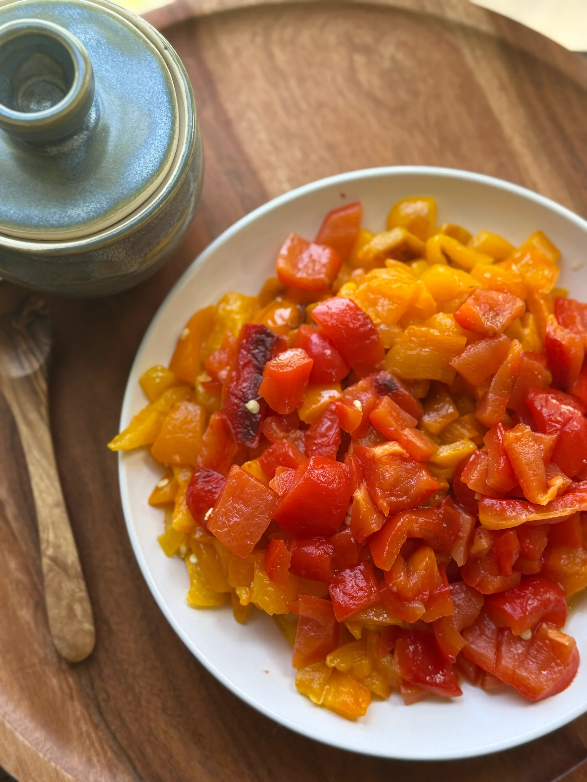 Roasted red and orange bell peppers arranged on a white plate. A wooden spoon and blue bowl on the side for decor. 