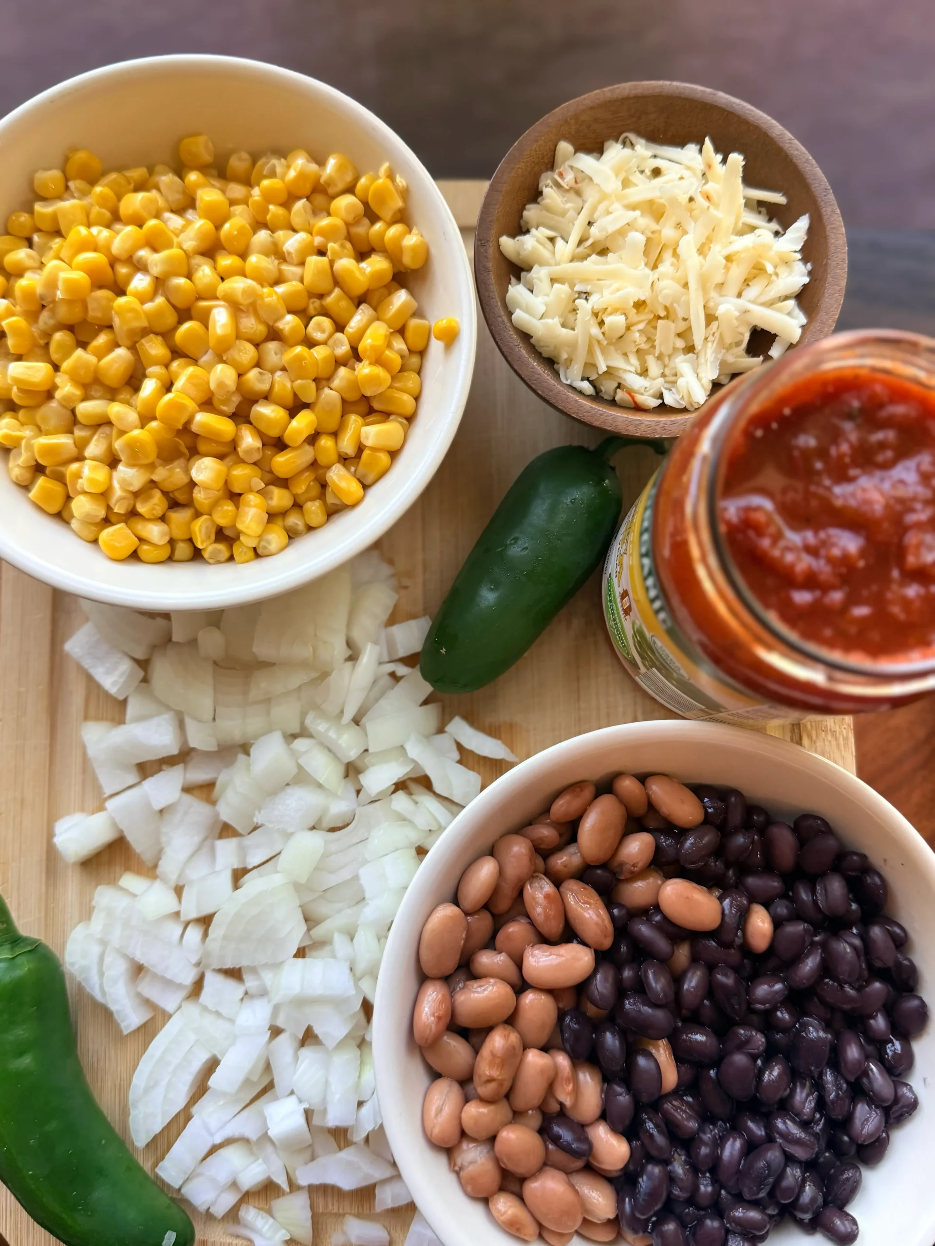 Cutting board with diced onions, 2 whole jalapenos, a jar of salsa, a bowl of cheese, a bowl of corn, and a bowl of black and pinto beans.