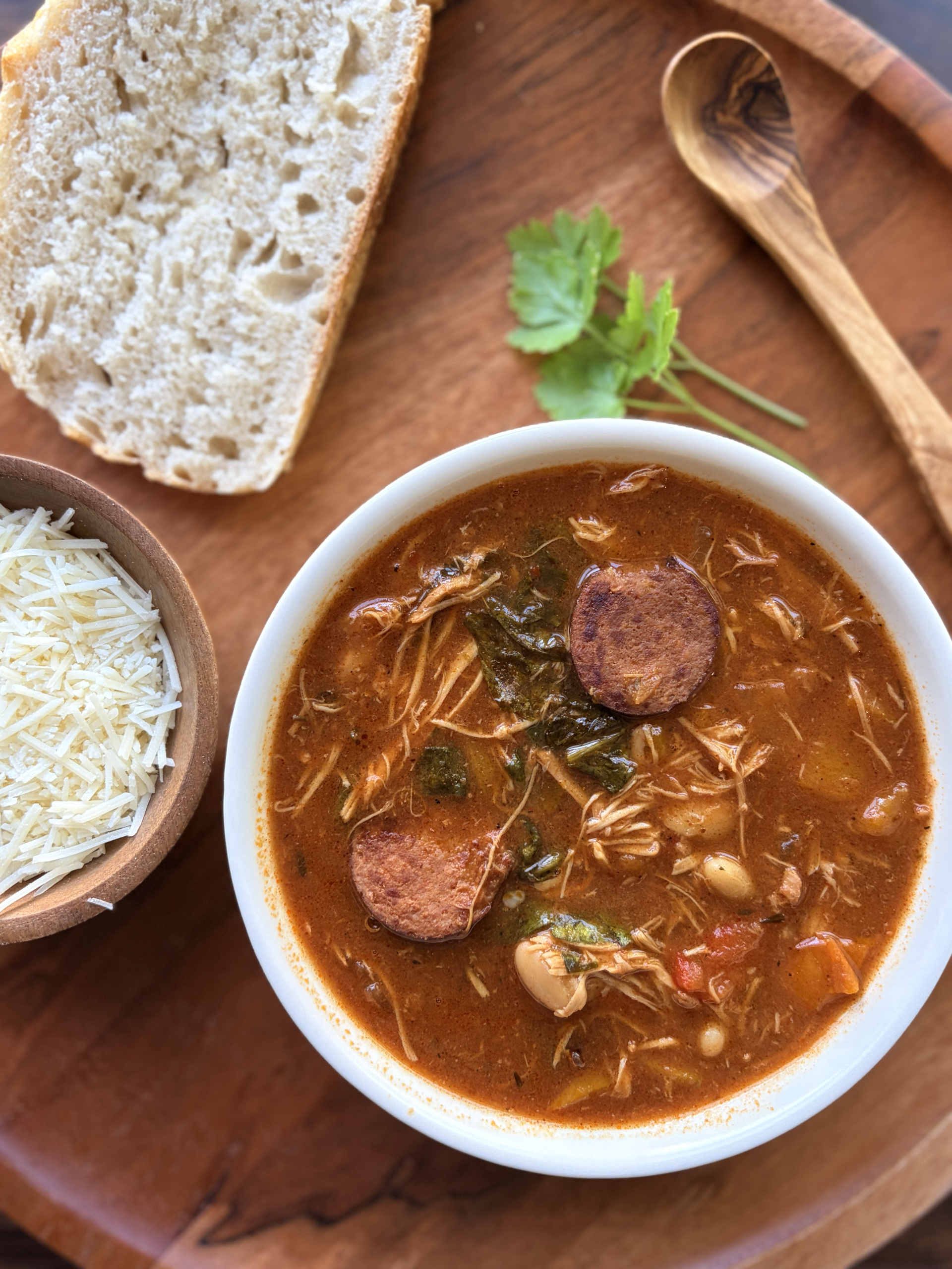 Tuscan sausage soup with shredded chicken in a bowl. A wooden bowl of shredded parmesan cheese and piece of sourdough bread on the side with a sprig of parsley for decor.