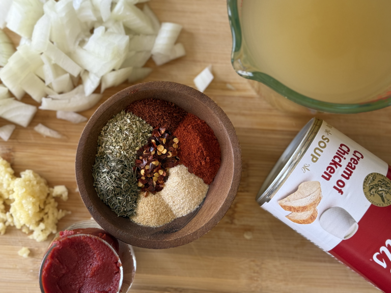 Blend of spices in a wooden bowl with a measuring cup full of chicken broth. A diced white onion, can of cream of chicken soup on the side with a diced white onion.