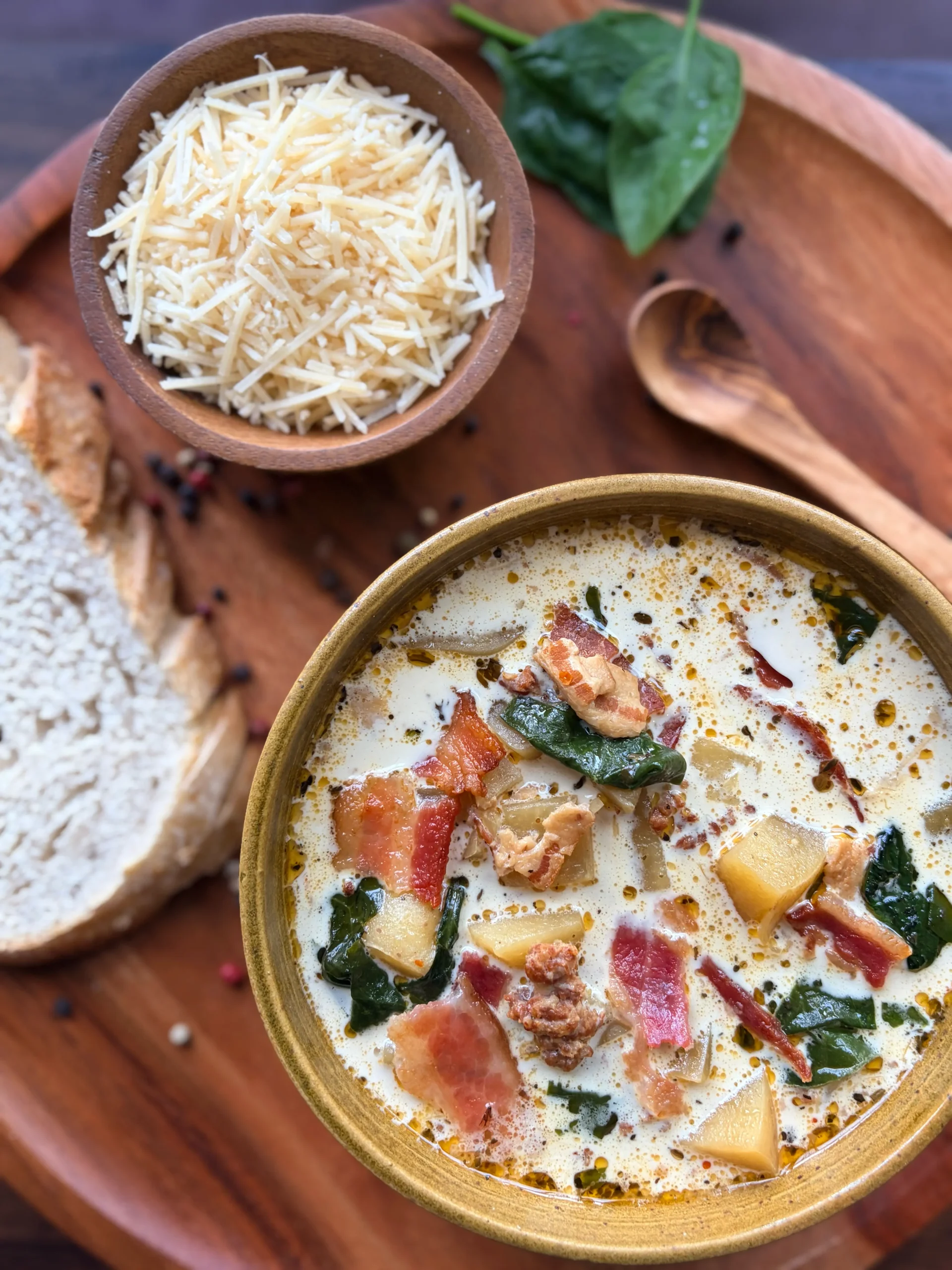 Bowl of soup with potatoes and bacon in a creamy broth. A slice of sourdough bread is on the side with a wooden spoon and bowl of grated parmesan cheese.