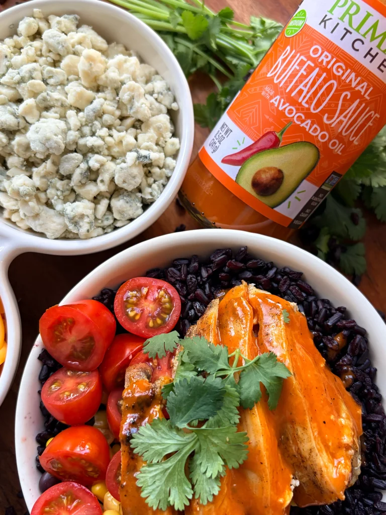 A bowl of buffalo chicken with cherry tomatoes and fresh cilantro. On the side is a bottle of Primal Kitchen Buffalo sauce with a bowl of blue cheese crumbles.