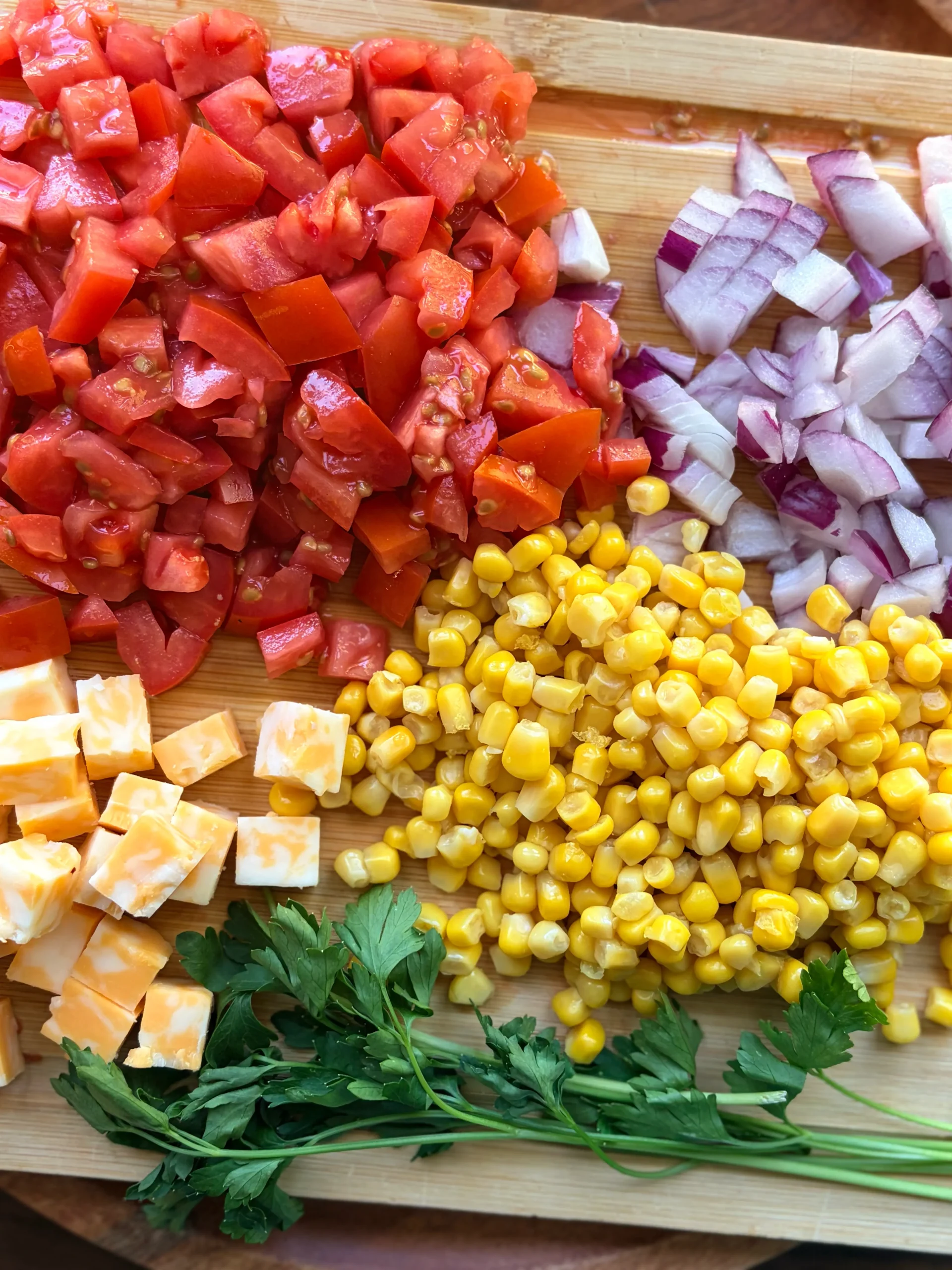 A cutting board with diced roma tomatoes, and diced red onion. There is corn and cheese and 3 sprigs of fresh parsley on the side.