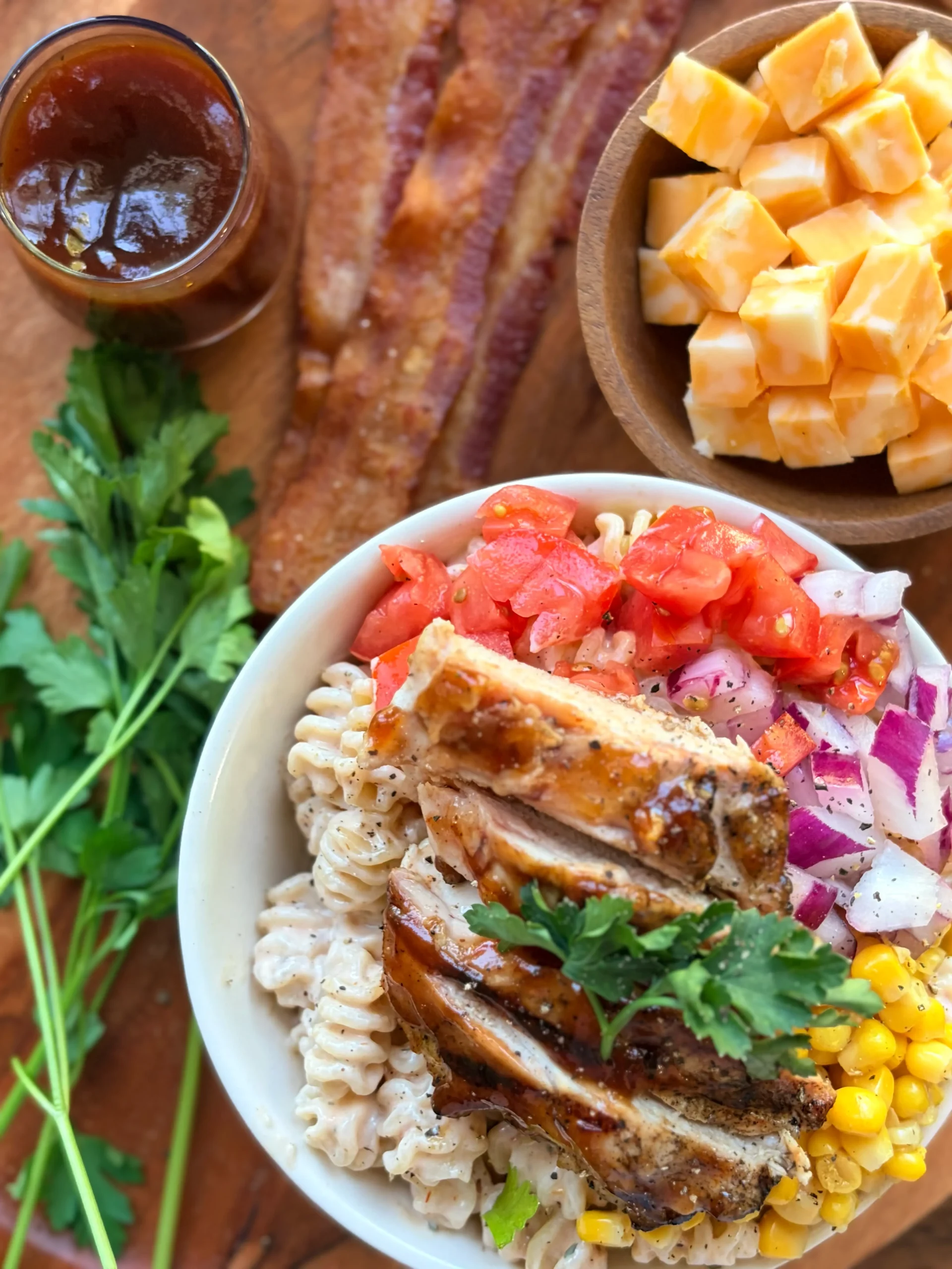 A bowl of barbecue chicken pasta salad. A bowl of cubed cheese is above it and there are 3 slices of bacon on the side.