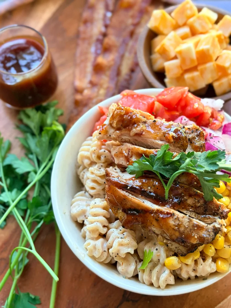 A bowl of barbecue chicken pasta salad. A bowl of cubed cheese is above it and there are 3 slices of bacon on the side.