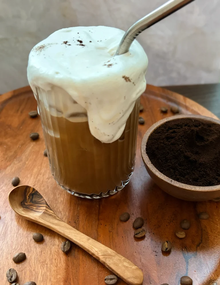 Iced coffee with coconut whipped cream, a bowl of ground coffee and a wooden spoon.