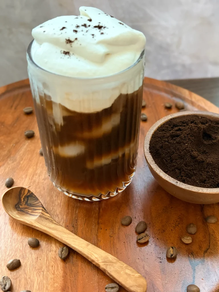 Iced coffee with coconut whipped cream, a bowl of ground coffee and a wooden spoon. 