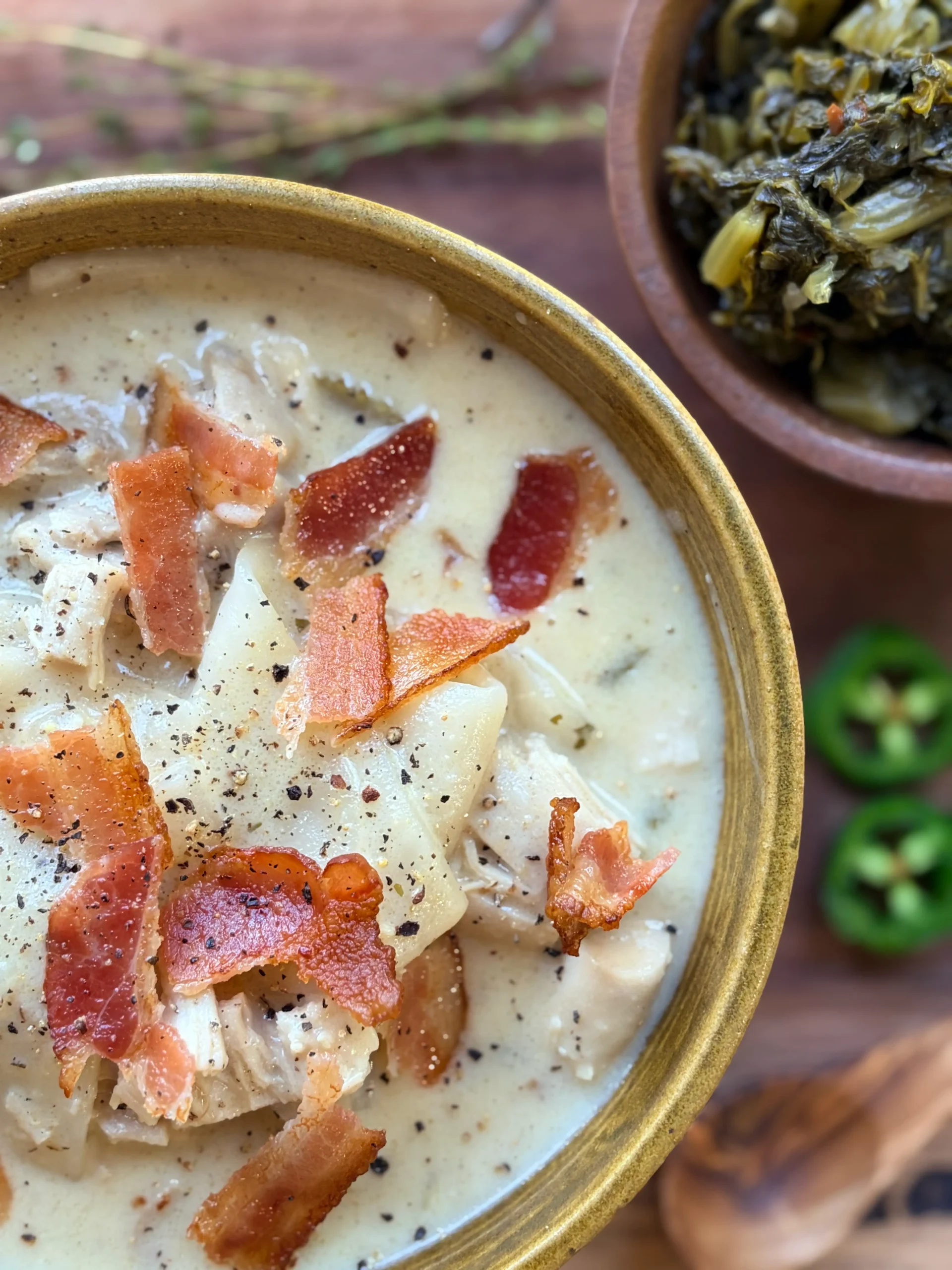 Bowl of chicken and dumplings topped with bacon. A side of turnip greens for decor.