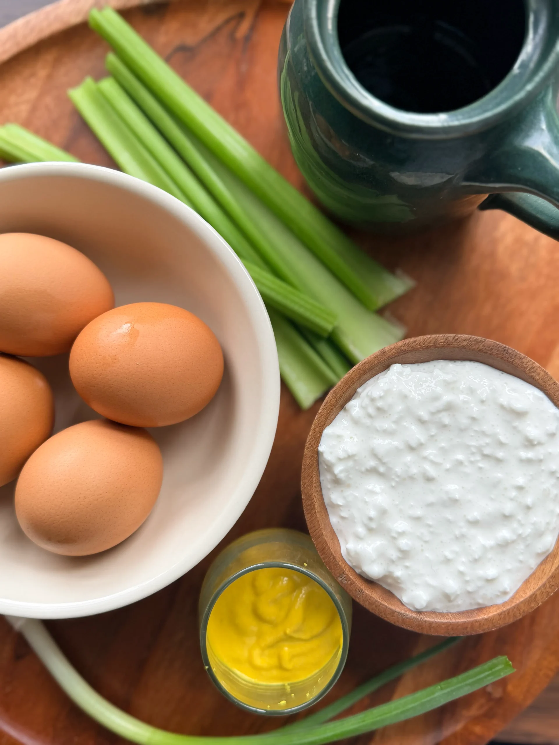 A bowl with 4 boiled eggs, a bowl with cottage cheese, a bowl with mustard. On the side are celery talks and 2 sprigs of green onion.