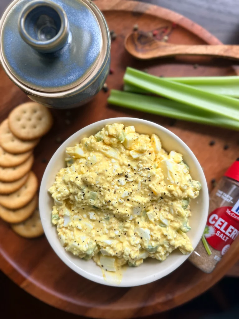 Bowl of egg salad with ritz crackers lined along the edge of the bowl. A few stalks of celery and some celery salt are on the side for decor.