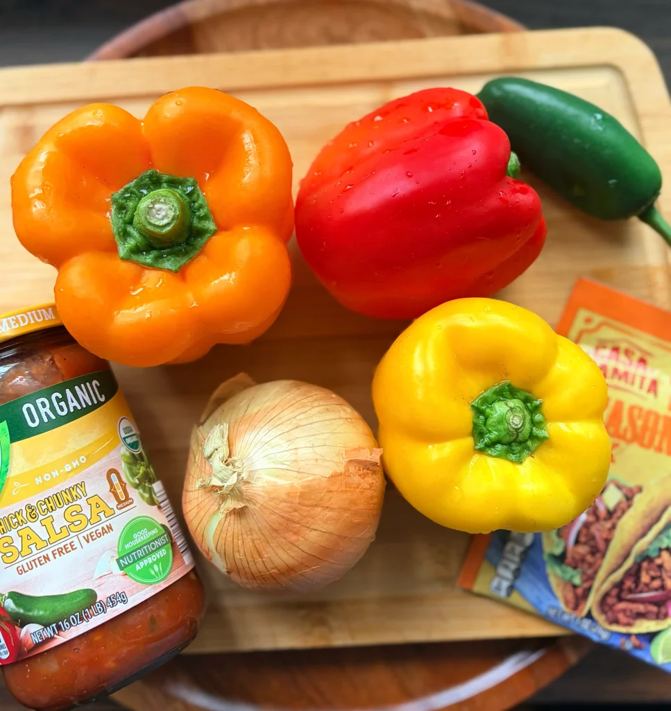 3 bell peppers, one onion, a packet of taco seasoning and a jar of unopened salsa on a wooden cutting board.
