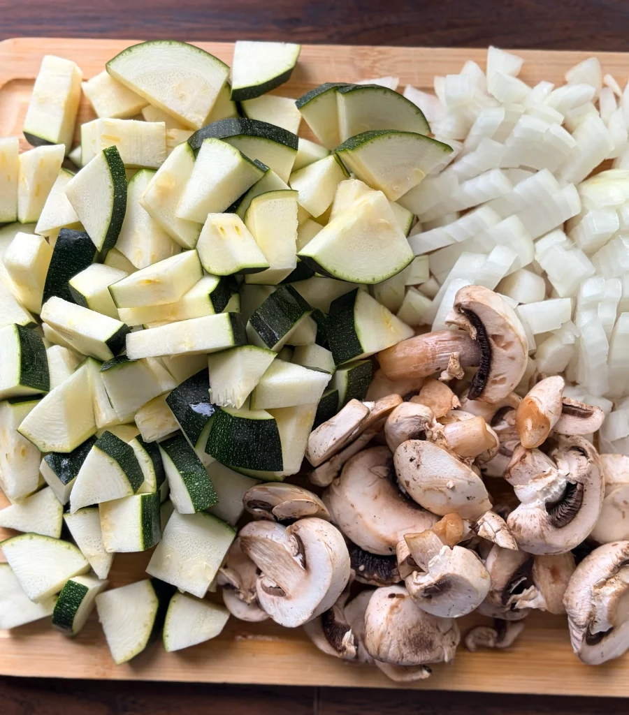 Diced white onion, diced zucchini, and sliced mushrooms all on a wooden cutting board. 
