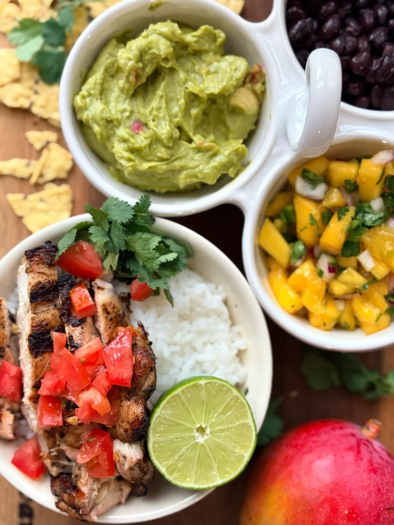 A bowl of jerk chicken and rice topped with tomato and a half of a fresh lime. In a serving dish there is an assortment of black beans, mango salsa, and guacamole.