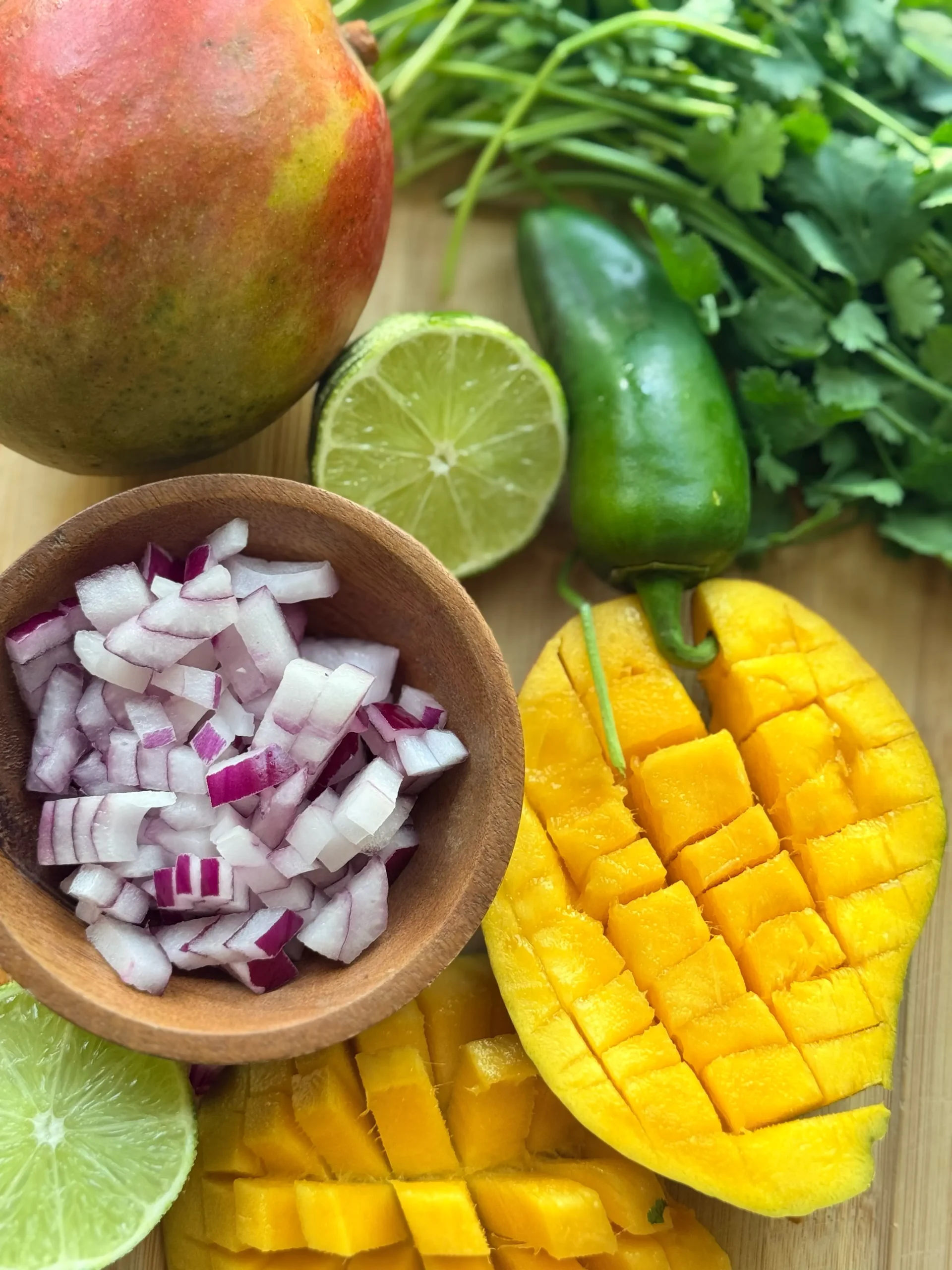 Ingredients to make a mango salsa, a cup of diced red onion and sliced mangoes. There is a whole mango and jalapeno sitting next to a bunch of fresh cilantro.