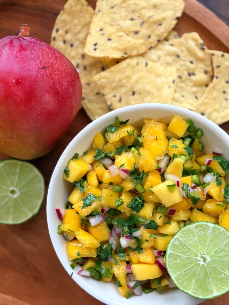 Bowl of mango salsa with a fresh lime. A few tortilla chips for decor and a whole mango sit next to the mango salsa.