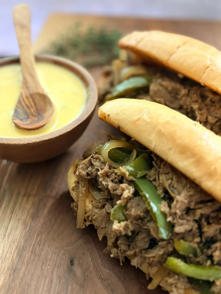 Two philly cheesesteaks that had no cheese on them yet. A bowl of cheese on the side with a wooden spoon.