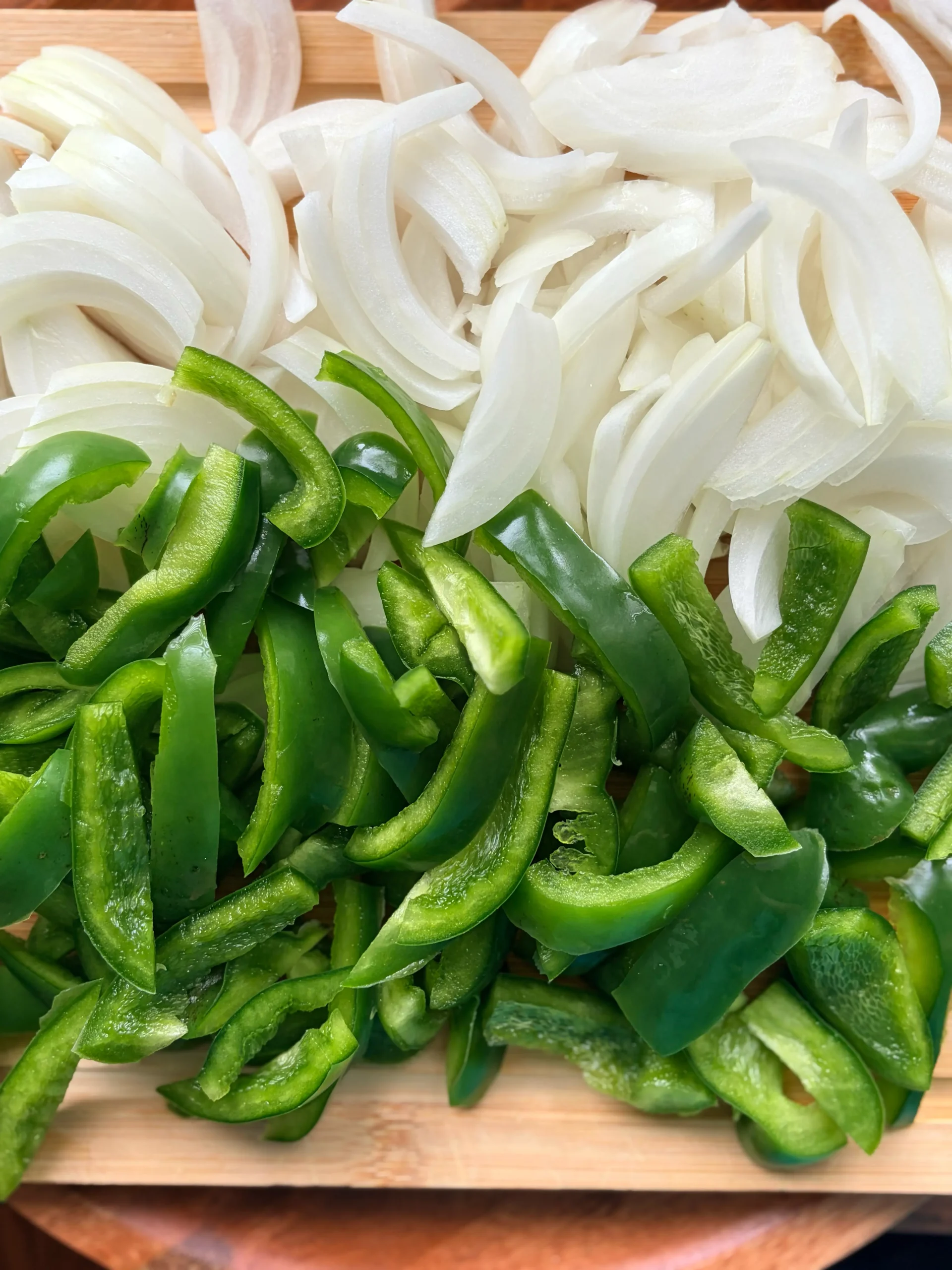 Sliced white onions next to sliced green bell peppers on a wooden cutting board.