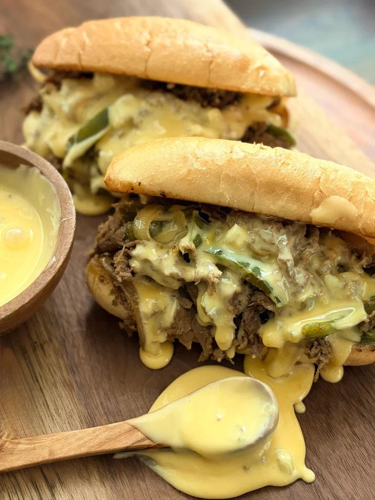Two philly cheese steaks with a bowl of cheese and a spoon to spread the cheese.