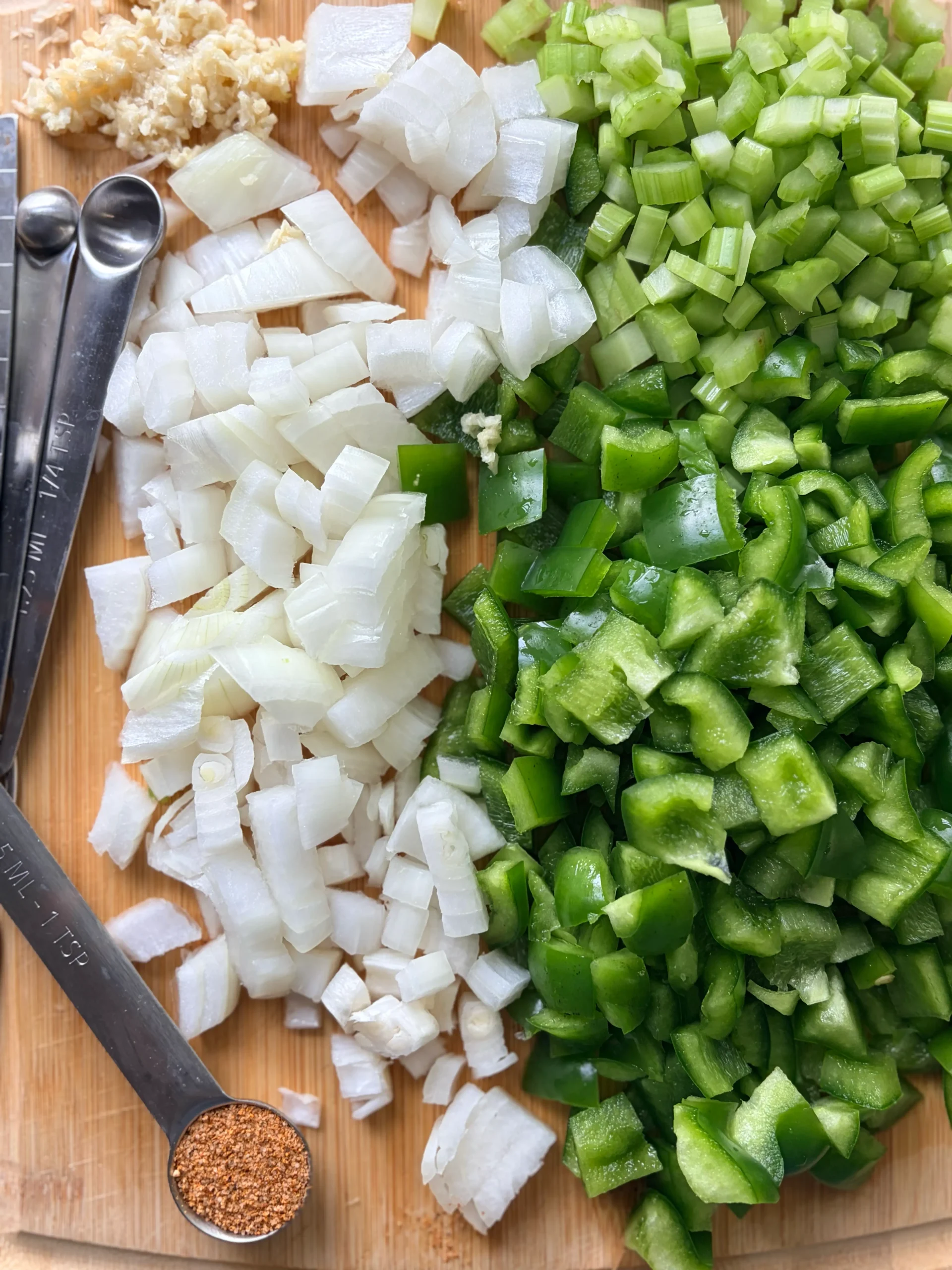 Minced garlic, white onion, bell pepper, and celery sit on a cutting board. There is a tsp of cajun seasoning still in the measuring spoon.
