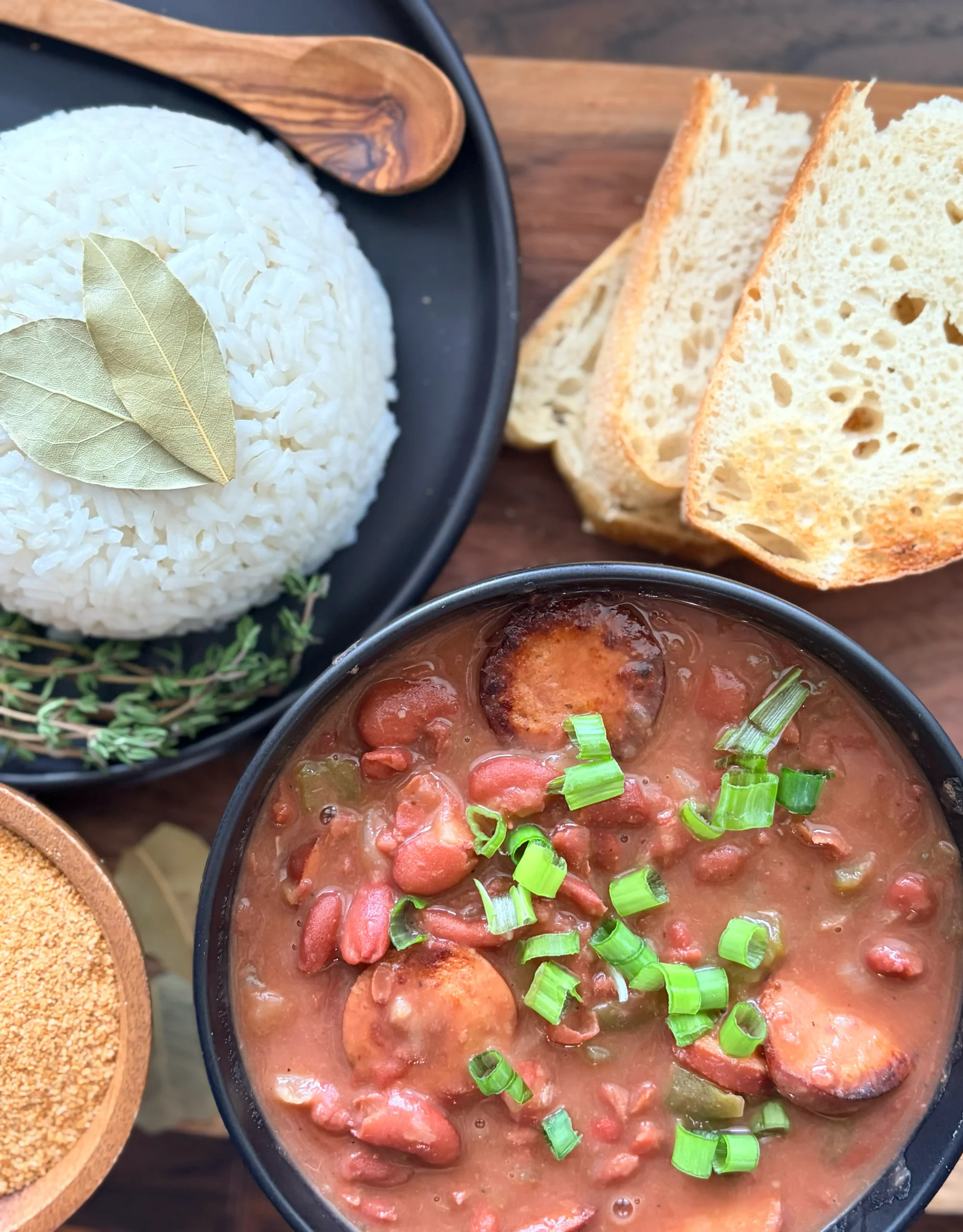 A bowl of red beans and rice with green onions sprinkled on top. There is a bowl of rice on the side with 3 pieces of fresh sourdough bread.